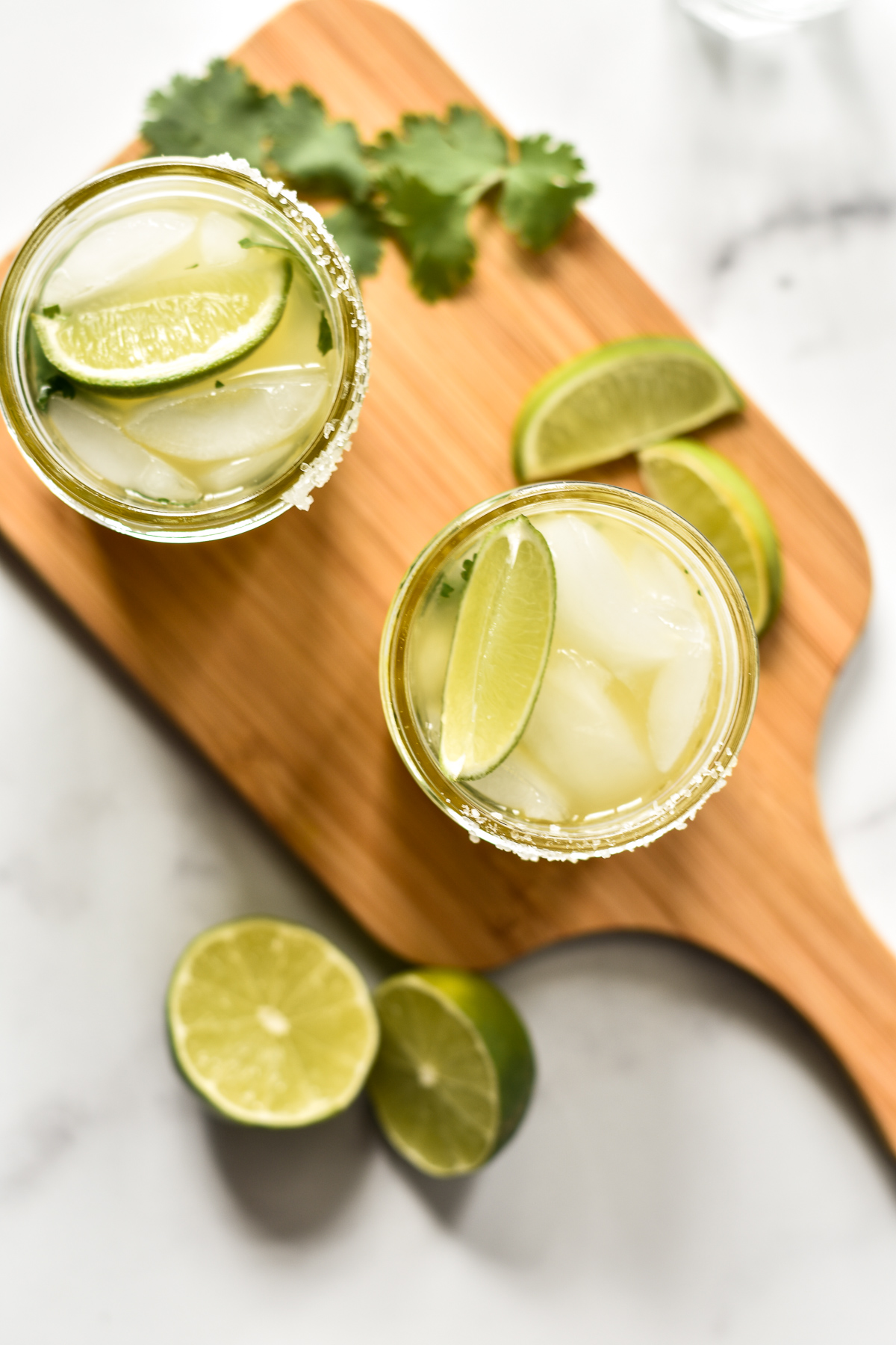 two pineapple cilantro margaritas on a wooden cutting board with limes and cilantro