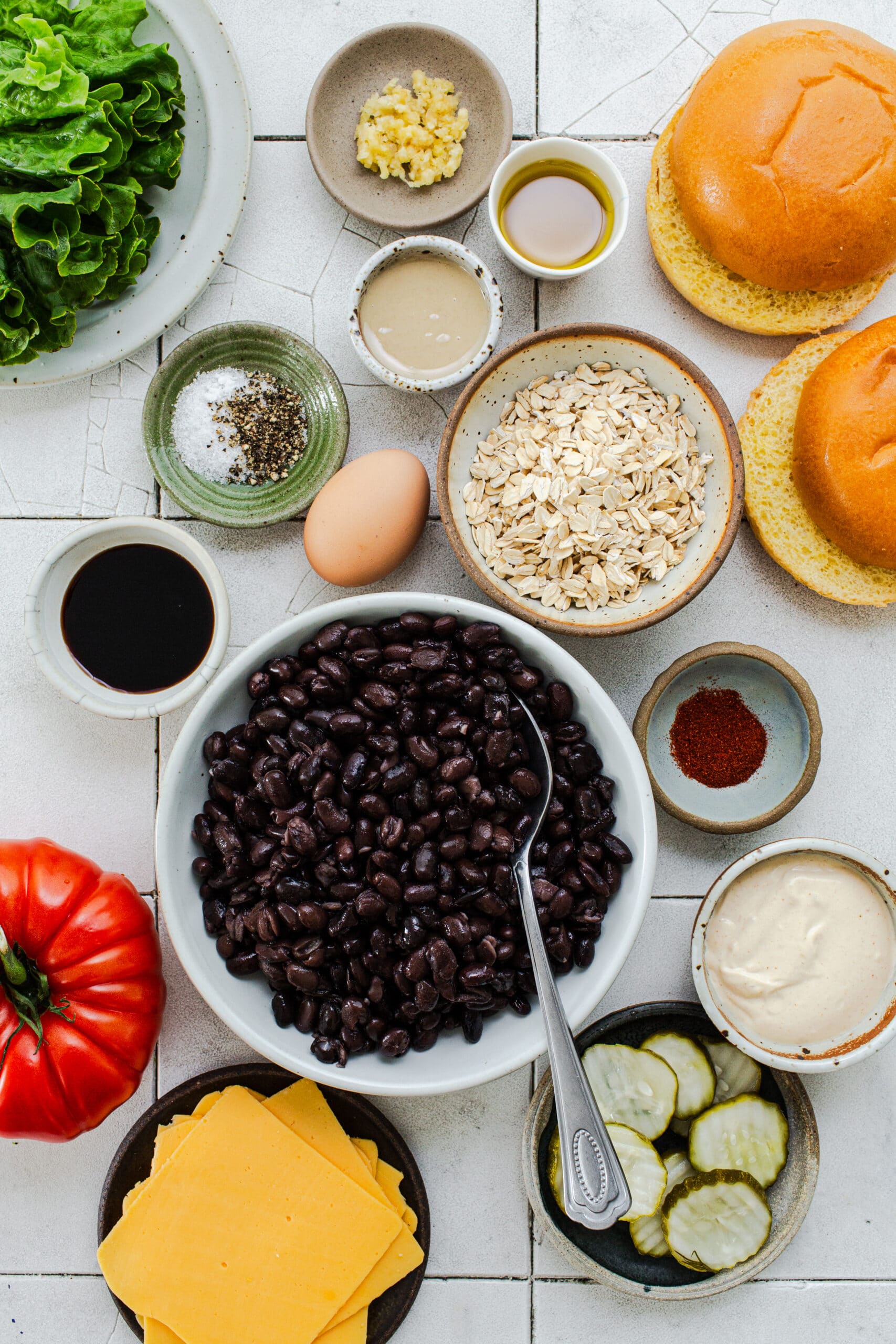 ingredients for black bean smash burgers in separate dishes including beans, oats, spices, egg, soy sauce, buns, cheese, tomatoes, pickles