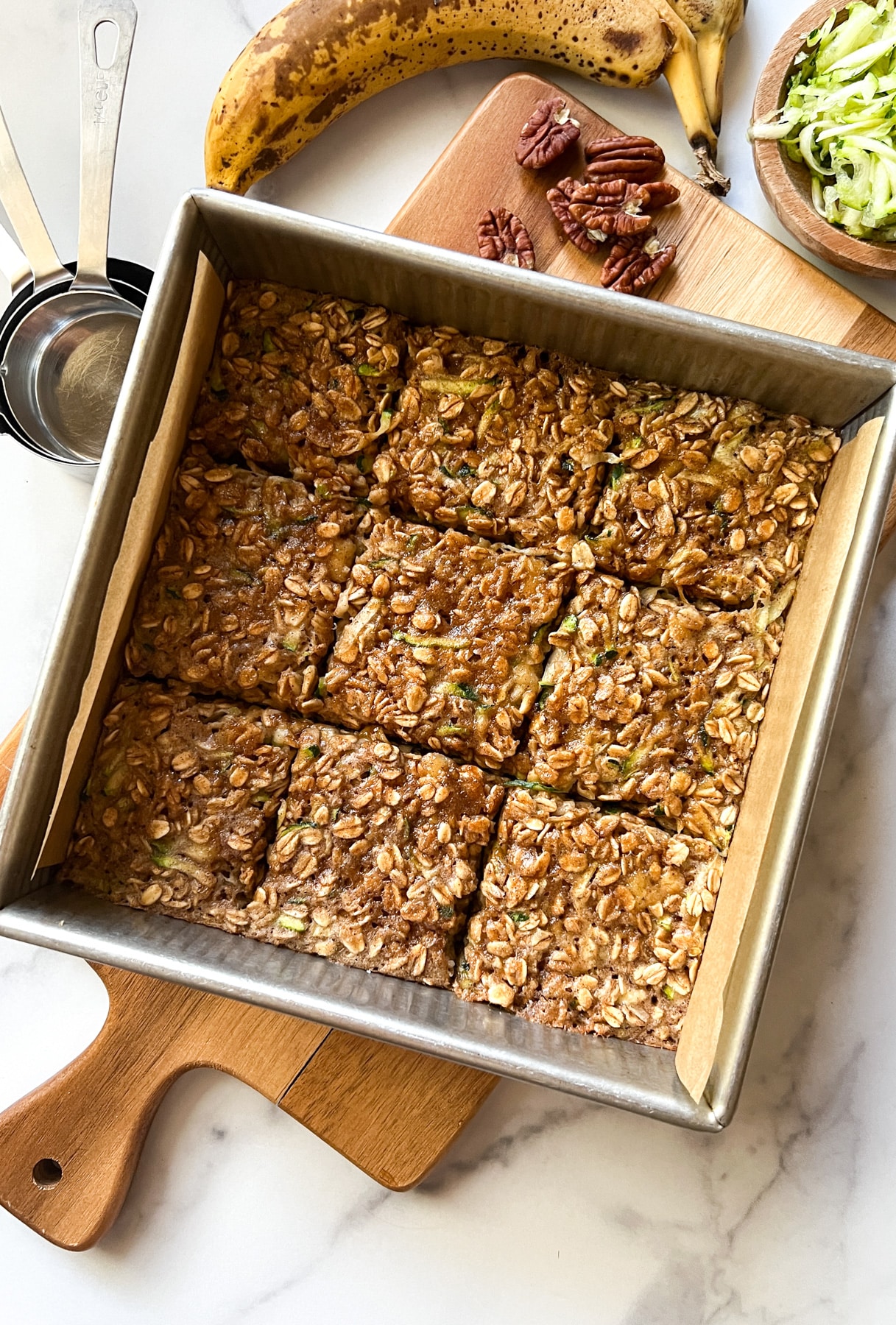 pan of baked oatmeal with measuring cups, banana, shredded zucchini and pecans on the side