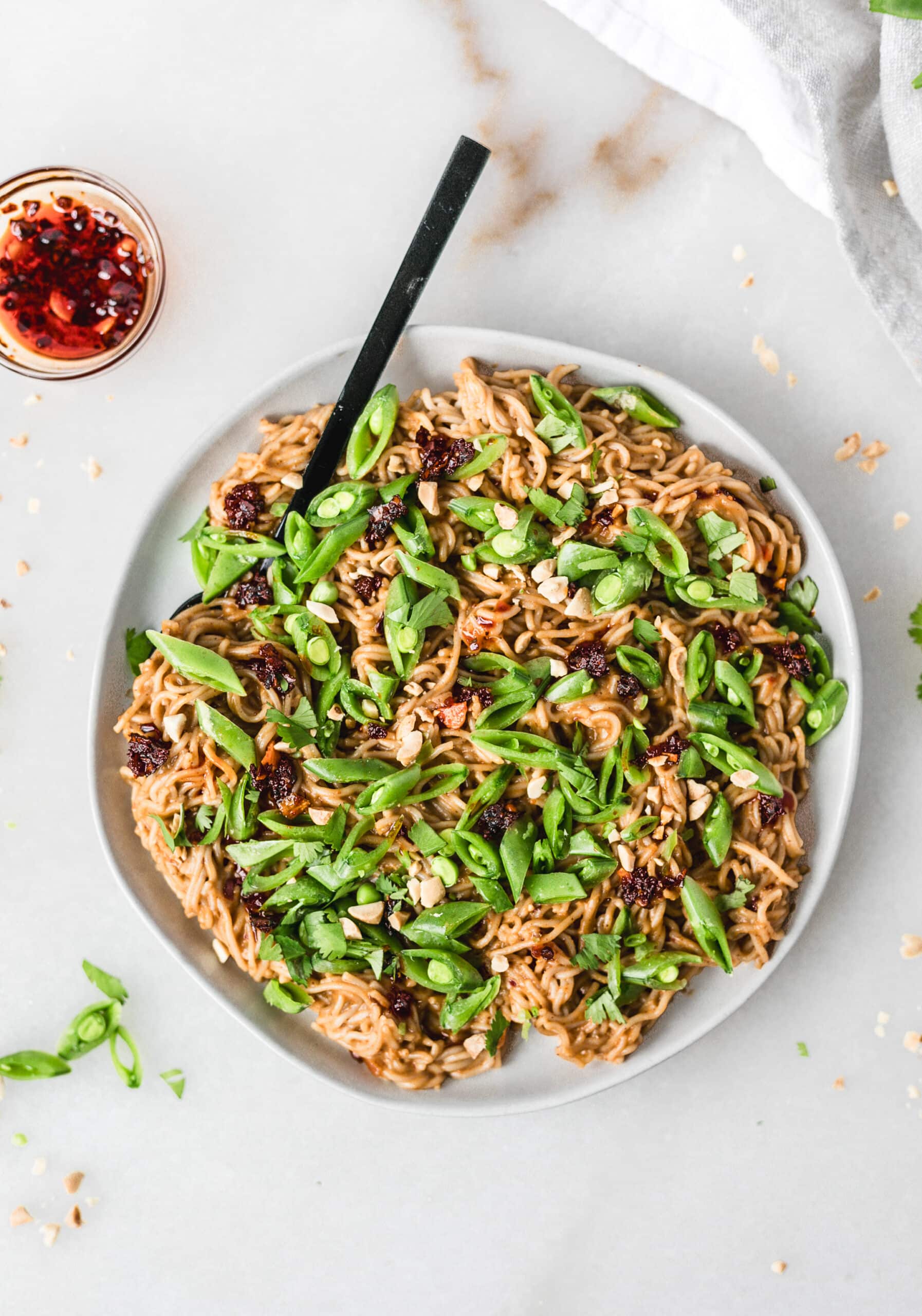 big plate of spicy peanut noodles with snap peas scattered on top and chili crunch on the side