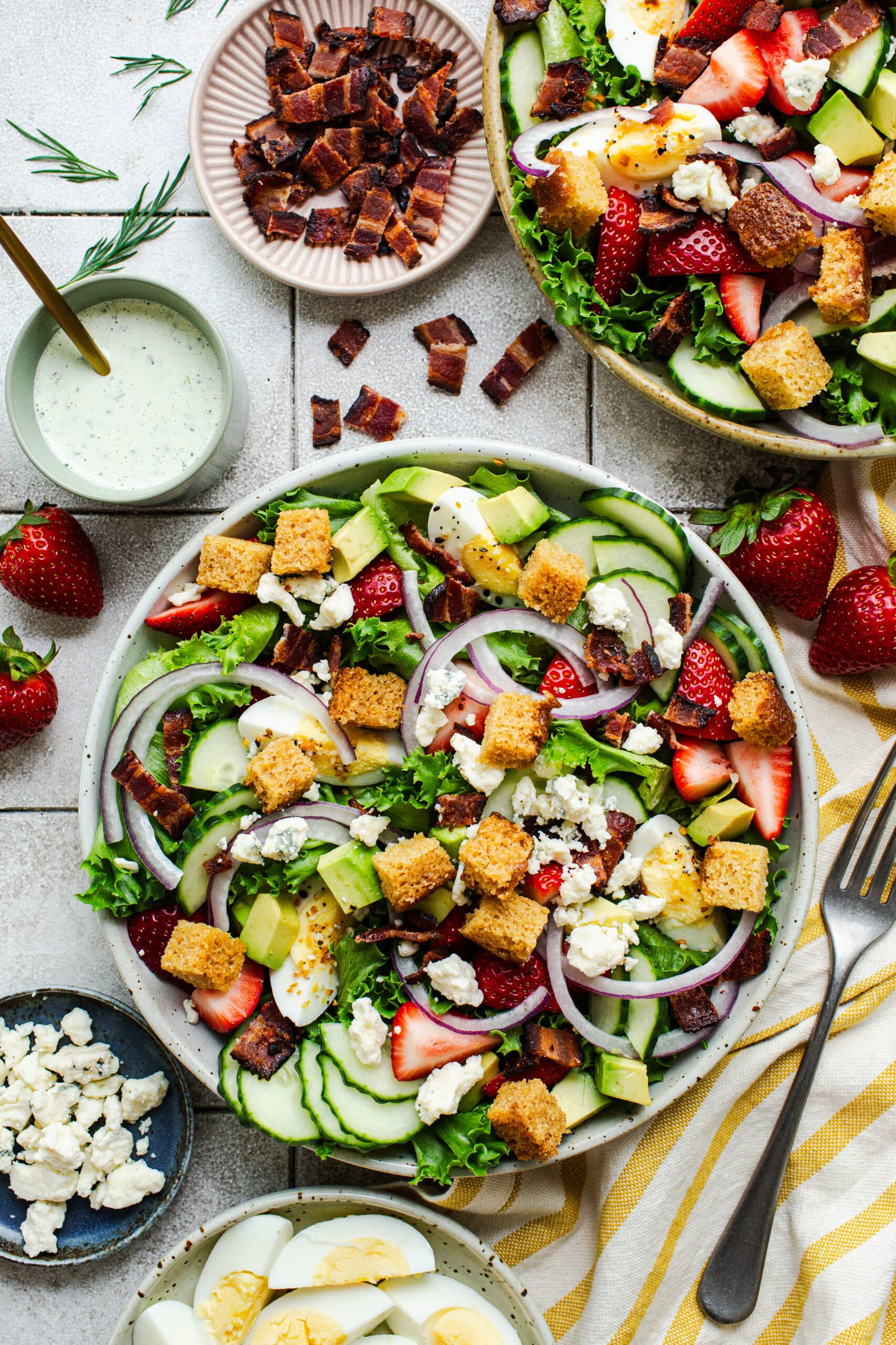 two plates of strawberry cobb salad with crispy bacon on the side and a cup of dressing