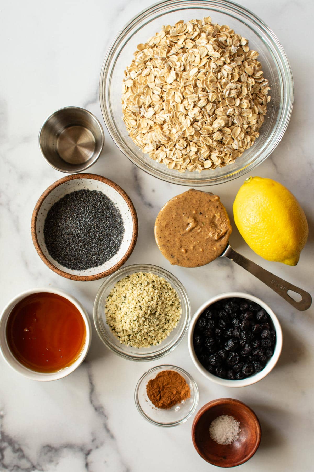ingredients in bowls including oats, almond extract, poppyseeds, almond butter, hemp hearts, dried blueberries, maple syrup, cinnamon, salt

