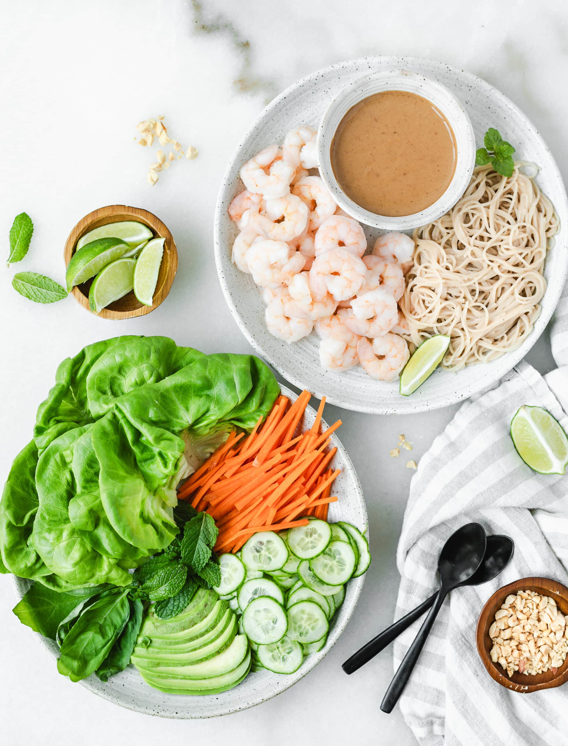 ingredients for shrimp lettuce wraps on plates including noodles, shrimp, lettuce, carrots, cucumber slices, avocado and peanut sauce in a dish