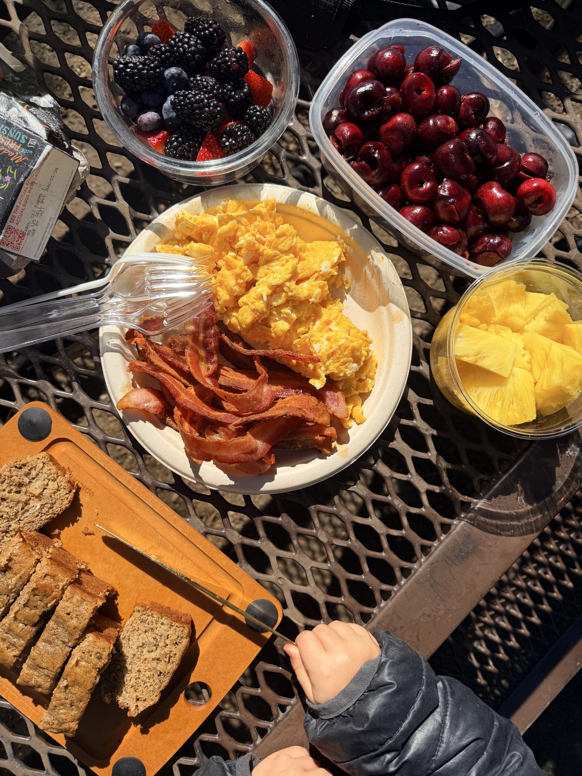 eggs and bacon on a plate, fruit and banana bread all on a camping table
